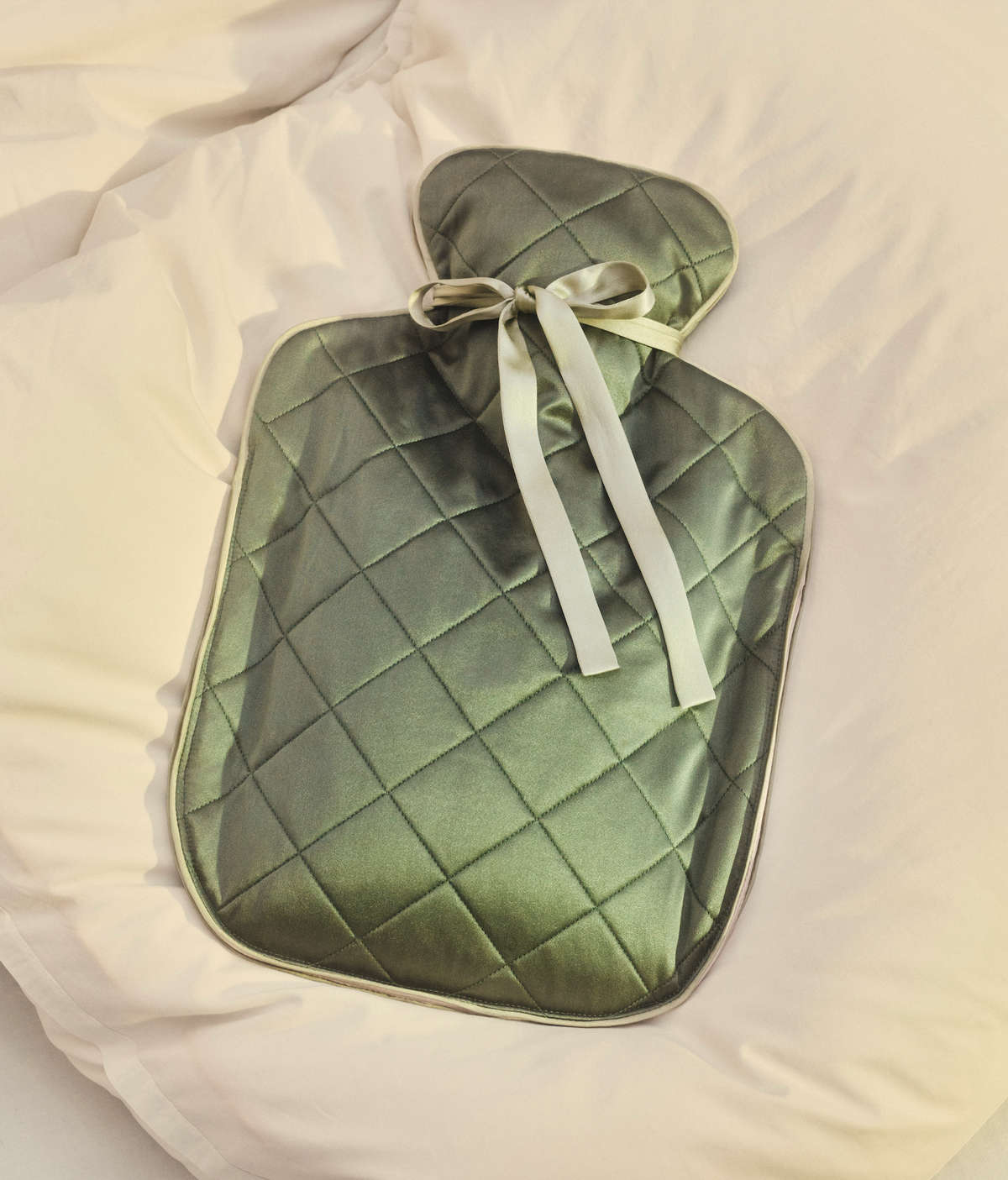 The Beauty Sleeper Large Hot Water Bottle in Meadow Bed, crafted in quilted green silk with a bow, resting on white bedding.